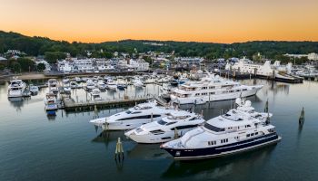 The image shows a marina with several luxurious yachts docked in calm waters during sunset, surrounded by a coastal town and green hills.