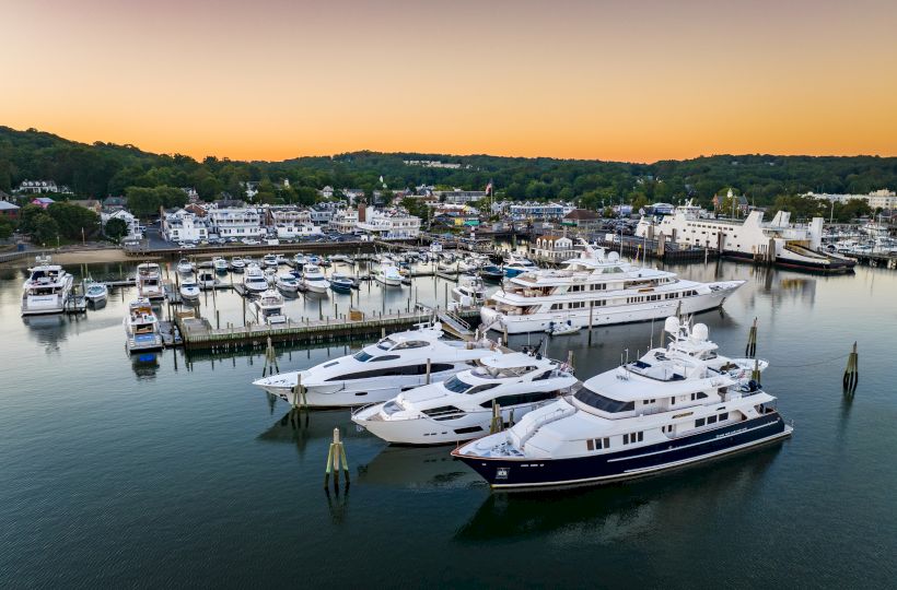 The image shows a marina with several luxurious yachts docked in calm waters during sunset, surrounded by a coastal town and green hills.