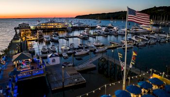 A marina at sunset with boats docked, an American flag flying, and people dining under blue umbrellas, creating a serene evening scene.